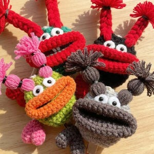 May include: A collection of handmade crochet monster toys in various colors. Each monster has large, expressive eyes, a wide mouth, and yarn hair. The color palette includes red, green, black, gray, orange, and pink.