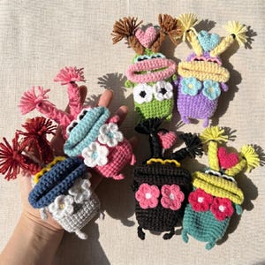 May include: A collection of colorful, handmade crochet frog-like creatures. Each has large eyes, a wide mouth, and decorative yarn accents. The frogs are in various colors, including green, pink, blue, purple, black, and yellow, with floral and heart details.