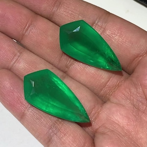 Puede incluir: Dos piedras preciosas de color verde vibrante en forma de pera con superficies facetadas. Cada piedra presenta un extremo puntiagudo y un borde superior más ancho y plano, mostrando una calidad translúcida y un color esmeralda brillante.