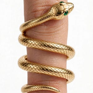 May include: A gold-toned ring shaped like a coiled snake with textured scales wrapping around a finger. The snake head features two small, vibrant green eyes, and the body forms a continuous spiral design that tapers to a pointed tail end.