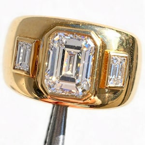 May include: A gold ring featuring a large, central emerald-cut clear gemstone set in a bezel, flanked by two smaller, rectangular emerald-cut clear gemstones on either side. The band is wide, polished, and smooth, with a high-shine metallic finish.
