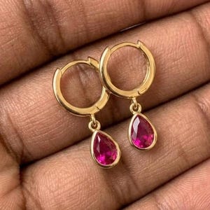 May include: A pair of gold hoop earrings featuring dangling teardrop-shaped fuchsia pink gemstones. Each earring consists of a circular gold hoop with a small, faceted, pear-cut stone suspended from the bottom, showcasing a vibrant pink hue.