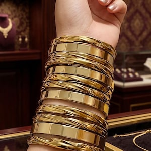 Pode incluir: Uma pilha de vários braceletes dourados usados no pulso. O conjunto consiste em uma alternância de faixas planas lisas e polidas com faixas texturizadas em estilo de corda torcida, todas com um acabamento metálico de alto brilho.