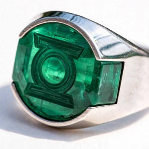 Puede incluir: Un anillo de tono plateado con una gran piedra preciosa circular de color verde translúcido. El centro de la piedra verde está grabado con el icónico logotipo circular de Green Lantern, que presenta un círculo central rodeado por un anillo y dos barras laterales.