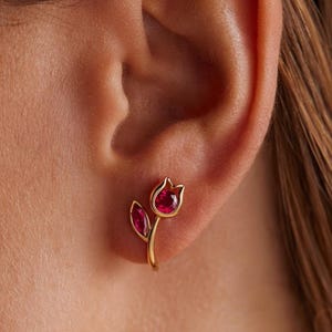 May include: A gold stud earring shaped like a tulip flower with a round, deep red gemstone forming the blossom and a marquise-cut red gemstone forming a leaf on the stem. The earring is secured to the earlobe with a post and backing mechanism.