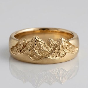 May include: A gold band ring featuring a textured, raised mountain range relief design that wraps around the exterior surface. The metal has a polished finish on the smooth lower portion and a detailed, rugged texture on the mountain peaks.
