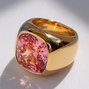 Op de afbeelding: Een gouden ring met een grote, kussengeslepen roze edelsteen in een gepolijste, dikke band. De binnenkant van de band is duidelijk gestempeld met de tekst 18K, wat de zuiverheid van het metaal aangeeft. Het ontwerp heeft een glad, afgerond profiel.