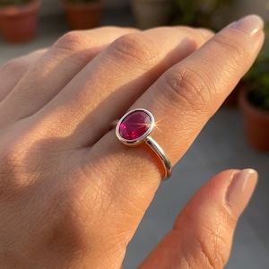 May include: A silver ring featuring a polished, oval-shaped, vibrant pinkish-red gemstone set in a smooth, bezel-style metal mounting. The band is plain and rounded, showcasing a clean, metallic finish that reflects the surrounding natural light.