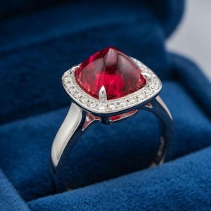 May include: A silver ring featuring a large, square-cut, deep red gemstone. The gemstone is encircled by a halo of small, clear stones. The ring is presented in a blue velvet box, accentuating its sparkle.