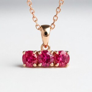 May include: A rose gold necklace with a pendant set with three round, bright red gemstones. The gemstones are arranged in a horizontal bar, suspended from a delicate chain. The necklace is shown against a soft, neutral background.