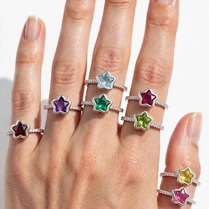 May include: A collection of silver-toned rings, each featuring a star-shaped gemstone in various colors including red, purple, light blue, green, dark pink, lime green, yellow, and pink, set on thin bands encrusted with small clear stones.