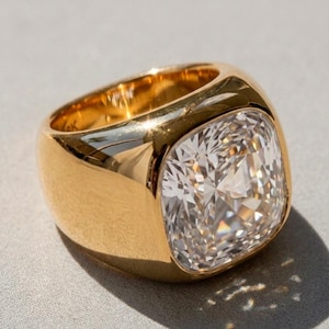 Può includere: Un anello in oro lucido con una grande pietra preziosa trasparente a taglio quadrato incastonata in una fascia spessa e liscia. La superficie metallica riflette la luce e la pietra presenta molteplici sfaccettature che catturano il bagliore.