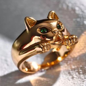 May include: A gold-toned ring featuring a sculpted cat design. The cat has two small, vibrant green gemstone eyes and its front paws are extended forward, resting on the top of the band. The metal surface is smooth, polished, and reflective.