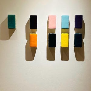 Könnte beinhalten: Ein abstraktes Kunstwerk mit neun rechteckigen Blöcken in verschiedenen Farben, darunter Türkis, Schwarz, Rosa, Hellblau, Gelb und Marineblau. Die Blöcke sind auf einer neutralen Wand angeordnet und werfen Schatten.