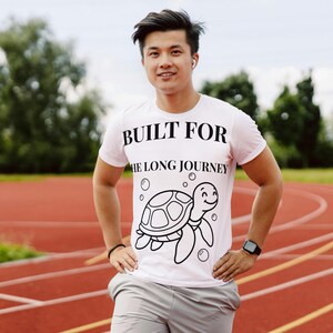 May include: A light pink t-shirt with the text "BUILT FOR THE LONG JOURNEY" and a cartoon turtle graphic. The shirt is worn by a person on a red running track.