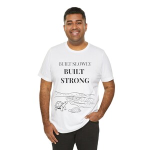 May include: White t-shirt featuring the text "BUILT SLOWLY BUILT STRONG" in black. Beneath the text is a black and white illustration of a beach scene with two turtles. The shirt is made of a soft, comfortable material.