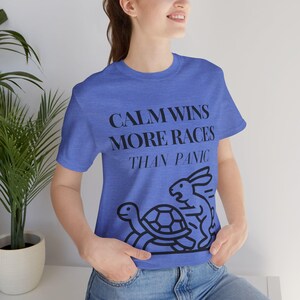 May include: A heather blue short-sleeve t-shirt featuring the black text CALM WINS MORE RACES THAN PANIC above a black line-art graphic of a turtle and a rabbit. The shirt has a crew neckline and a standard fit, showcasing a casual design.