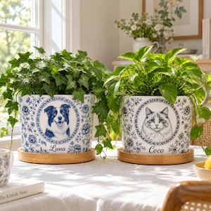 Op de afbeelding: Twee witte keramieken plantenpotten met blauwe bloemmotieven, elk op een ronde houten voet. De linkerpot toont een portret van een hond met de naam Luna, de rechterpot toont een kattenportret met de naam Coco.