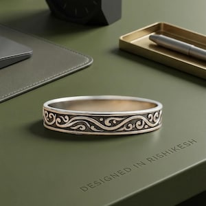 Puede incluir: Pulsera de metal plateado con un intrincado diseño de remolinos. La pulsera está sobre una superficie verde con el texto "DESIGNED IN RISHIKESH". Un reloj negro y una bandeja para bolígrafos están en el fondo.