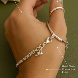 Può includere: Catena da mano in argento con un anello per il pollice e una catena a bracciale. La catena presenta un piccolo ciondolo e si collega a un anello per il pollice. L'anello per il pollice viene indossato sul pollice. La catena da mano è collegata a un braccialetto.