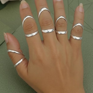 May include: A hand displaying several silver rings. The rings are of various designs, including curved and band styles. The jewelry is displayed against a neutral background, highlighting the metallic shine of the rings.