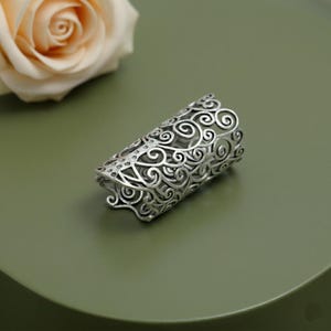 May include: A silver-colored finger armor ring with an intricate, openwork design of swirling patterns. The ring is cylindrical and covers a portion of the finger. A pale peach rose is in the background.