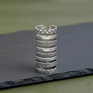 Puede incluir: Un anillo plateado con múltiples bandas, cada una con un patrón diferente. Los patrones incluyen triángulos, puntos y líneas. El anillo se muestra sobre una superficie gris oscuro.