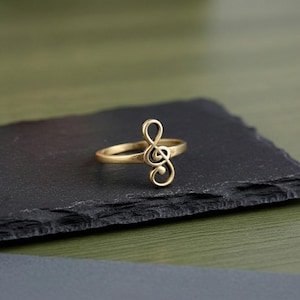 May include: A gold-colored ring featuring a treble clef design. The ring is set against a dark, textured surface, with a blurred green background. The ring's design is a stylized musical symbol.