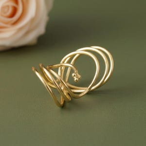 May include: A gold-colored ring with a unique, swirling design. The ring features multiple loops and a small, decorative element. The background is a solid, muted green, with a blurred peach-colored rose in the upper left corner.