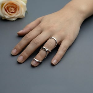 May include: A silver-toned metal finger splint ring featuring a smooth, polished finish. The design consists of two connected bands that wrap around the finger, with a central bridge piece that secures the structure across the middle joint.