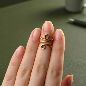 Peut inclure: Une bague réglable de couleur or avec un motif en spirale. La bague est portée sur un doigt, mettant en valeur son style unique et minimaliste. Le design de la bague présente deux boucles tourbillonnantes.