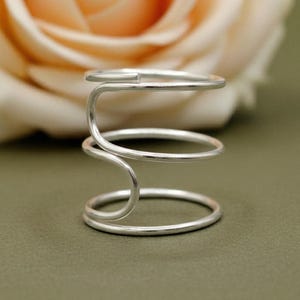 May include: A silver-colored ring holder with a unique, abstract design. The holder features three circular bands connected by a curved, flowing structure. The background includes a blurred peach-colored flower and a green surface.
