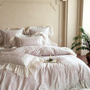 May include: A light pink bedding set featuring a delicate white lace pattern and ruffled edges. The set includes a duvet cover and multiple pillows, all adorned with intricate lace detailing and soft, pleated fabric borders for a layered look.