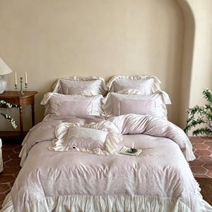 May include: A light lavender bedding set featuring a duvet cover and multiple pillows with white lace trim and ruffled edges. The duvet cover has a wide white ruffled skirt at the bottom and intricate white lace detailing along the top edge.