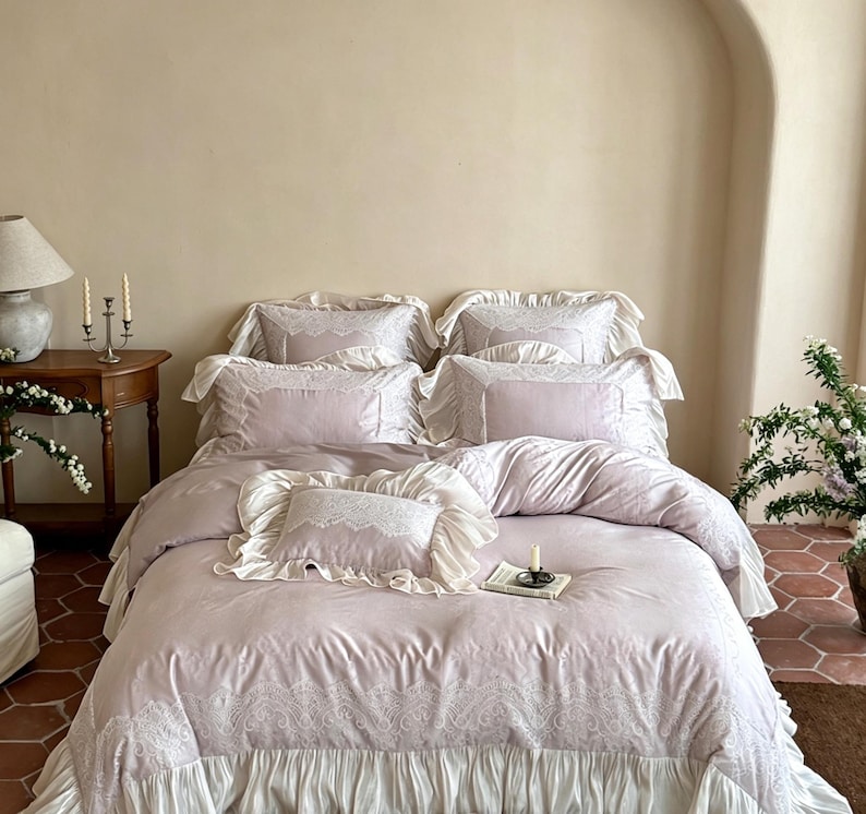 May include: A light lavender bedding set featuring a duvet cover and multiple pillows with white lace trim and ruffled edges. The duvet cover has a wide white ruffled skirt at the bottom and intricate white lace detailing along the top edge.