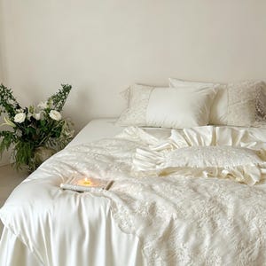 Puede incluir: Un juego de cama de color crema que incluye una funda n&oacute;rdica lisa con intrincados bordados de encaje, fundas de almohada a juego con paneles laterales de encaje y un coj&iacute;n decorativo cuadrado con borde de volantes sobre una cama.