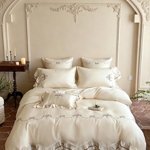 May include: A cream-colored bedding set featuring a duvet cover and pillowcases with delicate floral embroidery and ruffled edges. The set includes two large rectangular pillows, one smaller accent pillow, and a matching duvet cover.