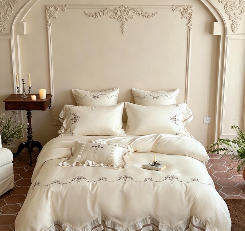 May include: A cream-colored bedding set featuring a duvet cover and pillowcases with delicate floral embroidery and ruffled edges. The set includes two large rectangular pillows, one smaller accent pillow, and a matching duvet cover.