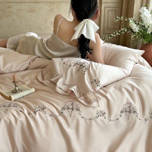 May include: A light beige bedding set featuring a delicate embroidered floral garland pattern across the duvet cover and pillowcase. The pillowcase is finished with a ruffled edge, and the fabric has a smooth, soft texture in a neutral tone.