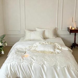 Puede incluir: Un juego de cama de color crema que incluye una funda n&oacute;rdica lisa, dos almohadas rectangulares con bordes de encaje y un coj&iacute;n cuadrado m&aacute;s peque&ntilde;o con borde de volantes. El conjunto incluye una manta texturizada y bordada.