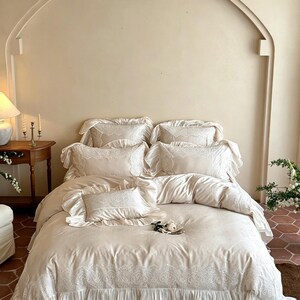May include: A cream-colored bedding set featuring a duvet cover and five pillows, all adorned with intricate white lace trim and ruffled edges. The fabric has a smooth, soft texture, and the lace detailing is present on the pillow borders.