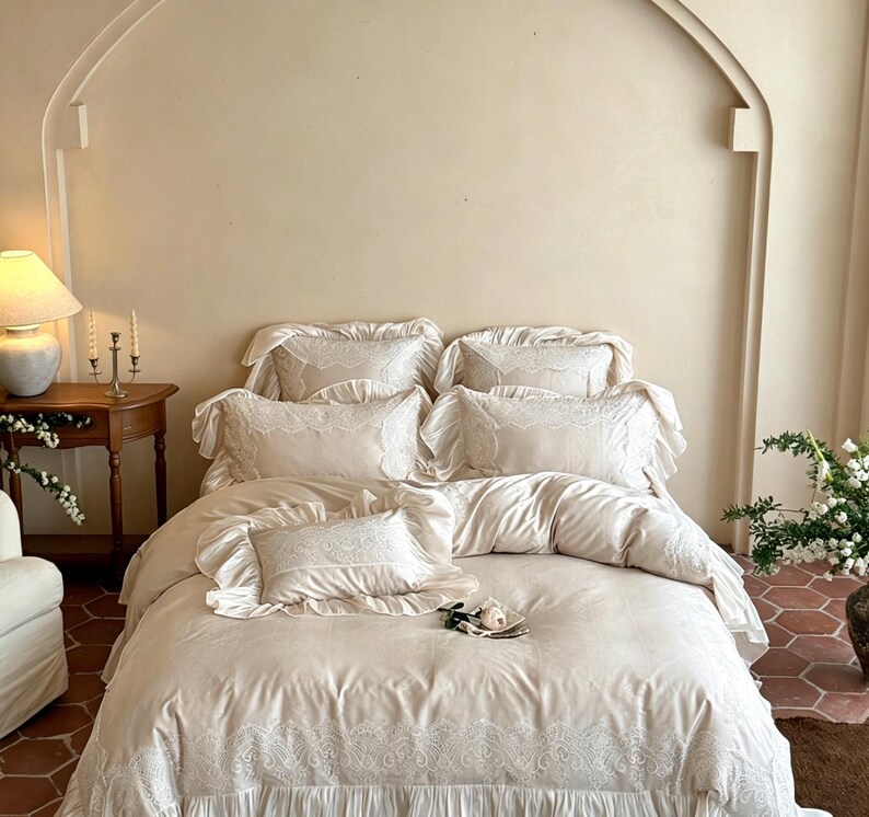May include: A cream-colored bedding set featuring a duvet cover and five pillows, all adorned with intricate white lace trim and ruffled edges. The fabric has a smooth, soft texture, and the lace detailing is present on the pillow borders.