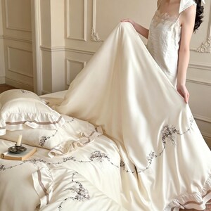 May include: A cream-colored bedding set featuring a duvet cover and pillowcases with ruffled edges, brown ribbon trim, and delicate floral embroidery. The fabric has a smooth, satin-like finish, and the set includes matching decorative accents.