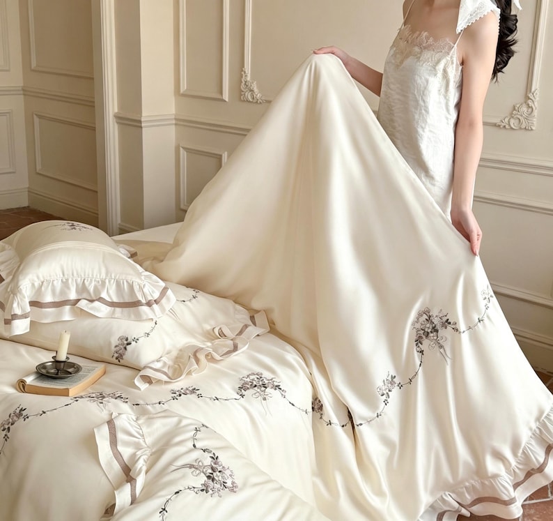 May include: A cream-colored bedding set featuring a duvet cover and pillowcases with ruffled edges, brown ribbon trim, and delicate floral embroidery. The fabric has a smooth, satin-like finish, and the set includes matching decorative accents.