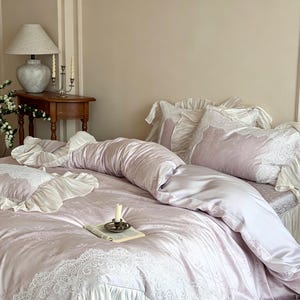 May include: A light lavender bedding set featuring a duvet cover and matching pillowcases with intricate white lace trim and ruffled edges. The fabric has a subtle tonal damask pattern, and the set is arranged on a bed with soft folds.