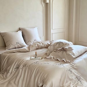May include: A beige bedding set featuring a duvet cover and two square pillowcases with delicate grey floral embroidery along the edges. The fabric has a smooth finish, and the pillowcases include ruffled trim details for a textured appearance.