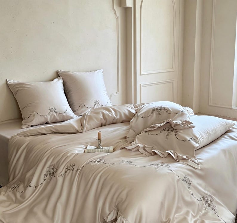May include: A beige bedding set featuring a duvet cover and two square pillowcases with delicate grey floral embroidery along the edges. The fabric has a smooth finish, and the pillowcases include ruffled trim details for a textured appearance.