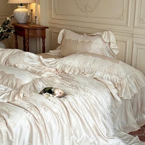 May include: A cream-colored bedding set featuring a duvet cover and matching pillowcases with intricate white lace trim and ruffled edges. The fabric has a subtle, repeating floral pattern and a smooth, satin-like finish, accented by a single rose.