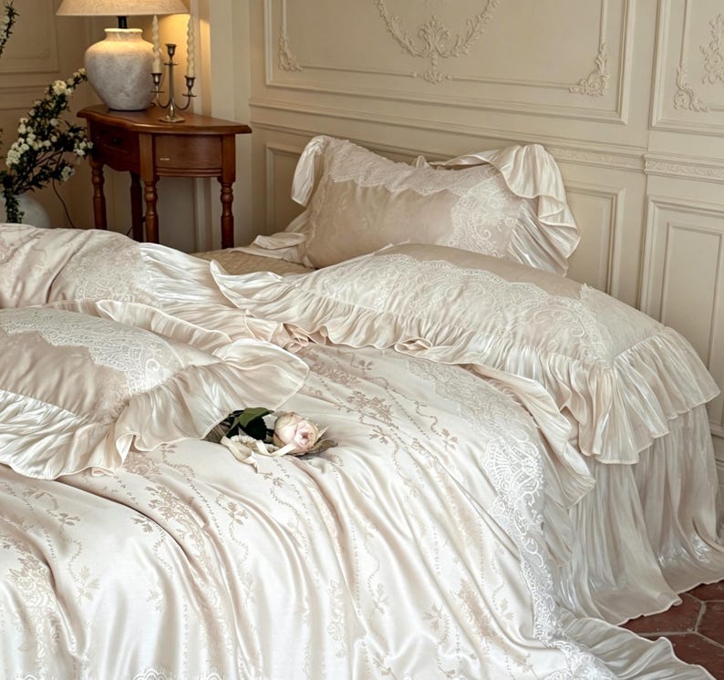 May include: A cream-colored bedding set featuring a duvet cover and matching pillowcases with intricate white lace trim and ruffled edges. The fabric has a subtle, repeating floral pattern and a smooth, satin-like finish, accented by a single rose.