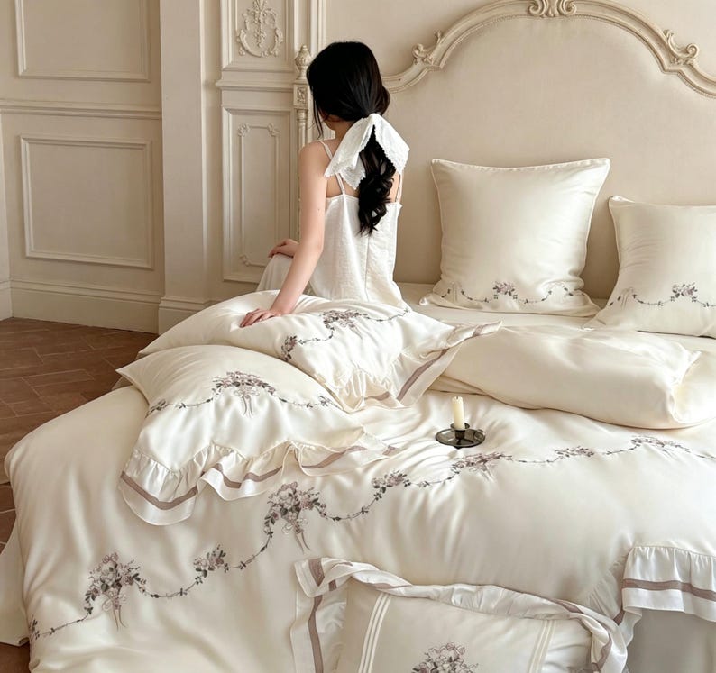 May include: A cream-colored bedding set featuring a duvet cover and multiple pillowcases with delicate floral embroidery and ruffled edges. The fabric has a smooth, satin-like finish, and the floral patterns are arranged in elegant swags.